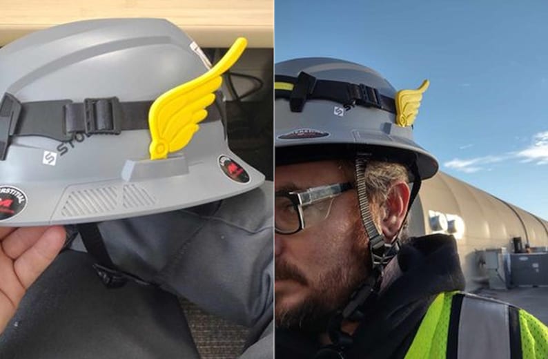May include: A gray hard hat with a yellow wing-shaped accessory attached to the side. The hard hat has a black strap and a white sticker with the word "STORM" on it.