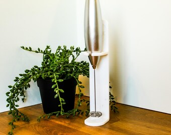 Könnte beinhalten: Ein silberner, handgeführter Milchaufschäumer mit dünnem Metall-Schneebesen, der in einem vertikalen weißen Kunststoffständer steht. Der Aufschäumer hat einen glatten, zylindrischen Metallkörper und einen kleinen runden Drahtbesen am Stab.