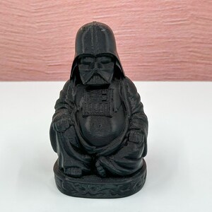 Puede incluir: Una figura negra de Buda con una m&aacute;scara de Darth Vader. La figura est&aacute; sentada en una postura meditativa con las manos juntas frente al pecho.