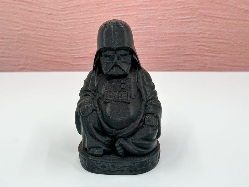 Puede incluir: Una figura negra de Buda con una m&aacute;scara de Darth Vader. La figura est&aacute; sentada en una postura meditativa con las manos juntas frente al pecho.