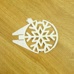 May include: A white plastic snowflake ornament with a circular design and a snowflake in the center. The ornament has two triangular extensions on one side and is laying on a wooden surface.