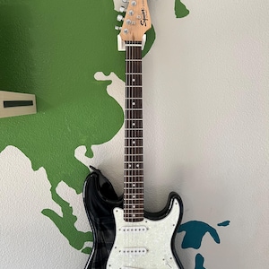 May include: A black electric guitar with a white pickguard and a white and black American flag sticker on the body. The guitar has a maple neck and a rosewood fingerboard. The guitar is hanging on a wall.
