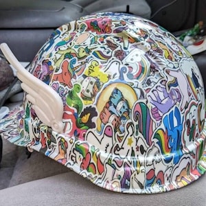 May include: A white hard hat covered in colorful unicorn and rainbow stickers. The hat has a white wing-shaped attachment on the side.