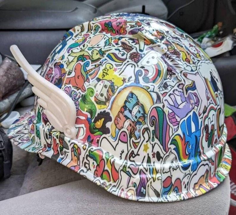 May include: A white hard hat covered in colorful unicorn and rainbow stickers. The hat has a white wing-shaped attachment on the side.