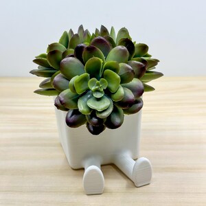 May include: A white ceramic planter shaped like a person sitting with legs crossed, holding a green and purple succulent plant.