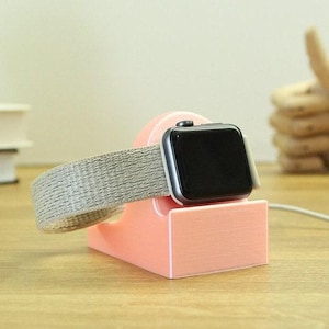 May include: A pink 3D printed charging stand for an Apple Watch with a gray fabric band. The watch is on the stand and the charging cable is plugged in.