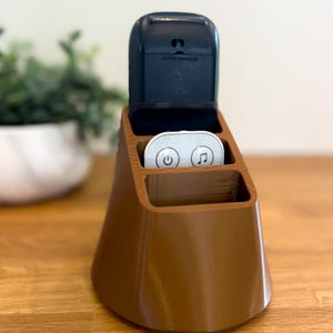 Può includere: Un organizzatore per telecomandi di forma conica marrone con tre fessure. Un telecomando nero è nella fessura superiore e due telecomandi bianchi sono nelle fessure inferiori. L'organizzatore è su una superficie di legno.