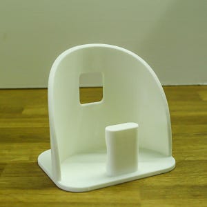 Puede incluir: Soporte de teléfono de plástico blanco con la parte posterior curvada y un recorte para un teléfono. El soporte tiene una pequeña base rectangular.