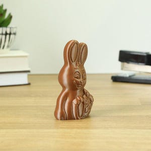 Puede incluir: Un conejito de chocolate marrón que sostiene una cesta de huevos. El conejito tiene una cara sonriente y está de pie sobre una superficie de madera.