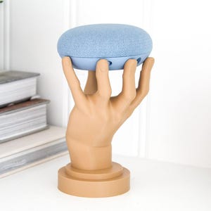 May include: A light blue Google Home Mini smart speaker is held by a brown hand sculpture. The sculpture is on a round base.