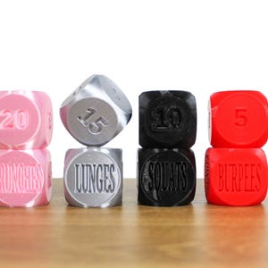 May include: Four sets of stacked dice with numbers and exercise names. The top dice are pink, silver, black, and red. The bottom dice are pink, silver, black, and red. The numbers on the top dice are 20, 15, 10, and 5. The exercise names on the bottom dice are "Crunches", "Lunges", "Squats", and "Burpees".