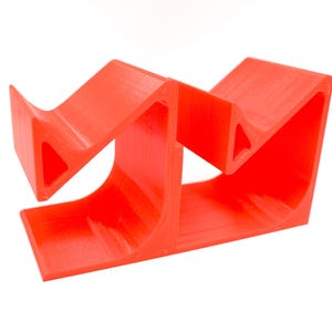 May include: A bright red, 3D-printed phone stand. The stand has a unique, geometric design with curved supports and angled surfaces. The stand is designed to hold a phone upright for hands-free use.