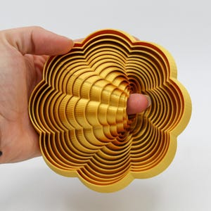 May include: A gold-colored metal flower-shaped object with multiple layers. The layers are stacked on top of each other, creating a three-dimensional effect.