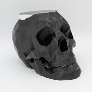 May include: A black, geometric skull-shaped decorative object. The skull has a faceted design and a hollow interior. The top of the skull has a flat, open surface. The object is made of a matte black material.
