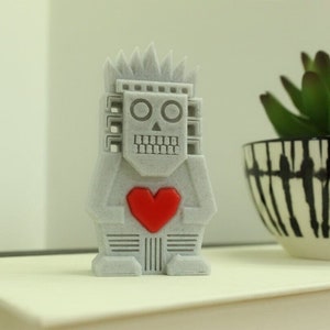 May include: A gray, geometric robot figurine with a red heart on its chest. The robot has a stylized face with circular eyes and a toothy grin. It has a spiky crown and a blocky design. The figurine is a decorative item.