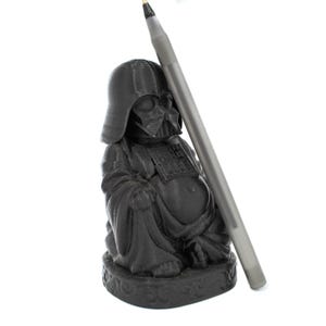 Puede incluir: Una figurita de pl&aacute;stico negro de Darth Vader vestido como Buda. La figurita est&aacute; sentada con las piernas cruzadas y tiene un bol&iacute;grafo plateado apoyado en ella.
