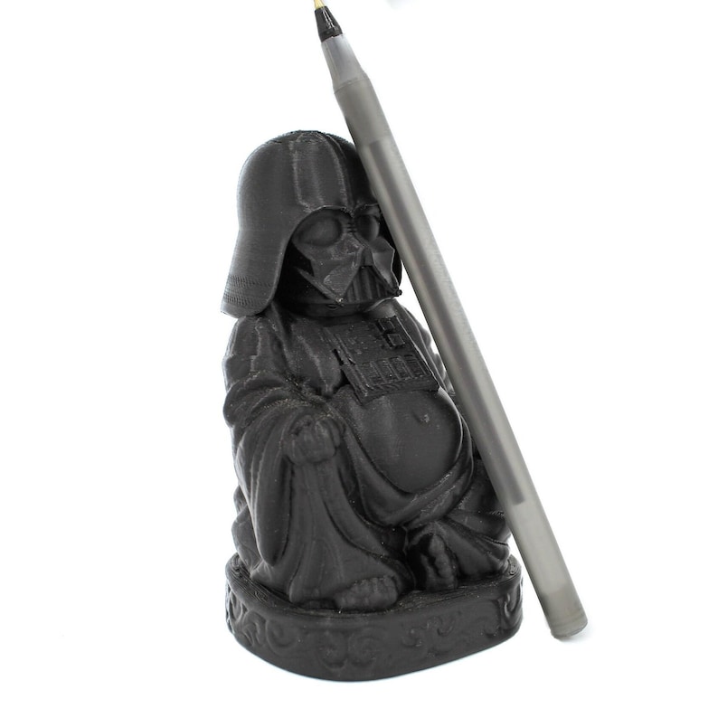 Puede incluir: Una figurita de pl&aacute;stico negro de Darth Vader vestido como Buda. La figurita est&aacute; sentada con las piernas cruzadas y tiene un bol&iacute;grafo plateado apoyado en ella.