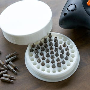 May include: A white plastic container with a lid, filled with a variety of small screwdriver bits. The container is open and the bits are visible. There are also some loose bits scattered on a wooden surface.