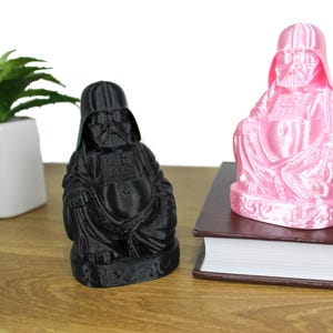 Puede incluir: Dos figuras de Darth Vader vestidas como Buda, una negra y otra rosa. La figura rosa est&aacute; sobre un libro marr&oacute;n. La figura negra est&aacute; sobre una superficie de madera.