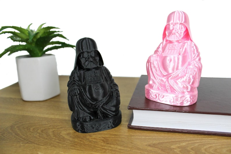 Puede incluir: Dos figuras de Darth Vader vestidas como Buda, una negra y otra rosa. La figura rosa est&aacute; sobre un libro marr&oacute;n. La figura negra est&aacute; sobre una superficie de madera.