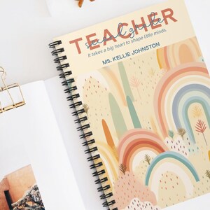 May include: A spiral-bound notebook with a rainbow design. The cover reads "TEACHER" and the quote "It takes a big heart to shape little minds." The notebook is open on a white surface with sunglasses and a paper clip.