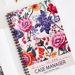May include: A spiral bound notebook with a floral design featuring pink, red, purple, and yellow flowers. The cover has the text "Annie Matthews Case Manager Genius, Rockstar, Legend".