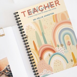 May include: Spiral-bound notebook with a rainbow design. The cover reads "TEACHER" and the quote "It takes a big heart to shape little minds." The notebook is open on a white surface with sunglasses and a paper clip.