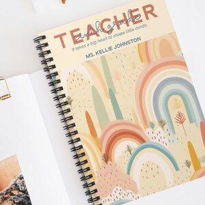 May include: A spiral-bound notebook with a rainbow design. The cover reads "TEACHER" and the quote "It takes a big heart to shape little minds." The notebook is open, showing blank pages. A gold paper clip and sunglasses are also visible.