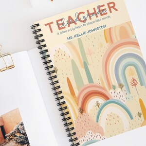 May include: Spiral-bound notebook with a rainbow design. The cover reads "Teacher" and "It takes a big heart to shape little minds. MS. KELLIE JOHNSTON." The notebook is open, showing blank pages. Sunglasses and a paper clip are also visible.