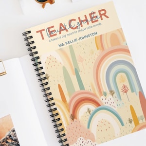May include: A spiral-bound notebook with a rainbow design. The cover reads "TEACHER" and the quote "It takes a big heart to shape little minds." The notebook is open on a white surface, with a gold paper clip and sunglasses nearby.