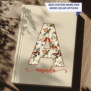 Op de afbeelding: Een crèmekleurig notitieboek met een groot bloemen "A" initiaalontwerp. De letter is gevuld met vlinders, bloemen en groen. De naam "Amanda" is eronder in een cursief lettertype geschreven. Tekst bovenaan zegt "VOEG EIGEN NAAM TOE EN MEER KLEUROPTIES".