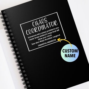 May include: Black spiral-bound notebook with the text "CHAOS COORDINATOR" in white. The notebook includes a quote and a space for a custom name. A gold binder clip and open book are in the background.