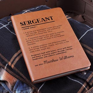 May include: A brown leather-bound notebook with the word "SERGEANT" in bold letters. The notebook is open, revealing text describing the qualities of a sergeant. The notebook rests on top of folded plaid shirts inside a suitcase.