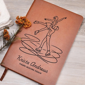 May include: A tan leather-like notebook featuring a black line art illustration of two figure skaters performing a lift. Below the illustration, the text reads "Krista Andrews" in a script font, followed by "FIGURE SKATING COACH" in block letters.