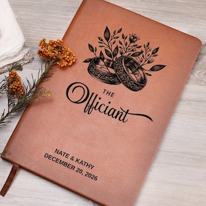 Pode incluir: Um caderno de couro sintético castanho com uma gravação preta de dois anéis entrelaçados rodeados por folhas. O texto diz "THE Officiant" acima de "NATE & KATHY" e "DECEMBER 20, 2026". Um ramo de flores cor de laranja está à esquerda do caderno.