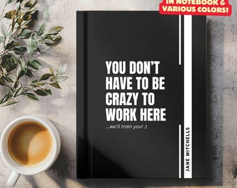 May include: A black notebook with the text "YOU DON'T HAVE TO BE CRAZY TO WORK HERE" in white. The notebook also has the text "...we'll train you! :)" and "JANE MITCHELLS". A coffee cup and plant are in the background.
