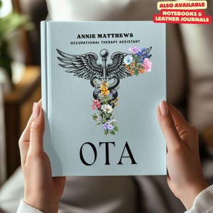Op de afbeelding: Een lichtblauwe notitieboekomslag met een zwart-wit medisch caduceus-symbool met bloemen en de tekst "ANNIE MATTHEWS OCCUPATIONAL THERAPY ASSISTANT OTA".