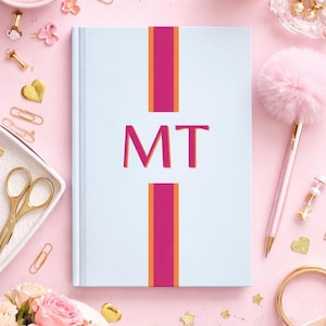 Op de afbeelding: Een lichtblauw notitieboek met de roze initialen "MT". Het notitieboek heeft een roze en oranje streepontwerp. De afbeelding is omgeven door roze accessoires, waaronder een pen, schaar en gouden briefpapier.