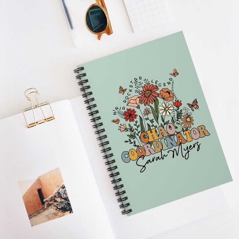 Op de afbeelding: Een spiraalnotitieblok met een lichtgroene omslag met een kleurrijk bloemenontwerp en de tekst "Chaos Coordinator Sarah Myers".
