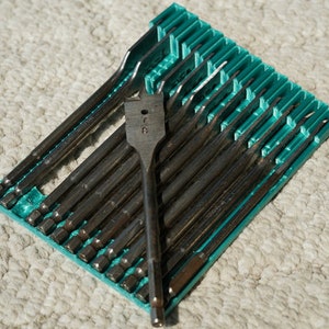 Puede incluir: Un juego de brocas de pala plateadas y negras en un soporte de plástico turquesa. Una broca se alza, mostrando su diseño plano en forma de paleta. Las brocas están ordenadas, sugiriendo un kit de herramientas para carpintería o construcción.