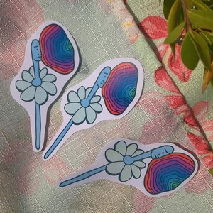May include: Three colorful stickers with a daisy and a face inside a rainbow swirl. The stickers are on a floral patterned surface.