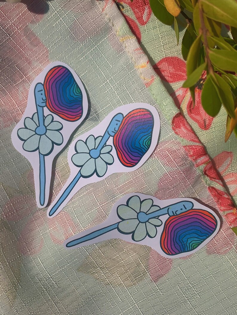 May include: Three colorful stickers with a daisy and a face inside a rainbow swirl. The stickers are on a floral patterned surface.