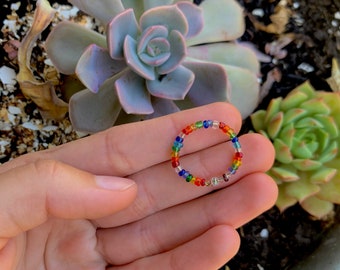 May include: A beaded ring with a rainbow color scheme, featuring red, orange, yellow, green, blue, and purple beads. The ring is held in a hand, with a succulent plant in the background.