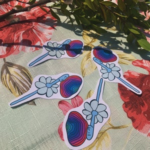 May include: Four colorful stickers featuring a psychedelic mushroom with a face and a daisy. The mushroom is a rainbow of colors, including red, orange, yellow, green, blue, and purple. The daisy is light blue.