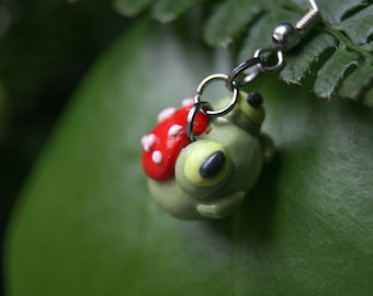 May include: A green frog with a red and white mushroom on its back is hanging from a silver chain. The frog has large yellow eyes and a small black nose. The frog is hanging against a green leafy background.