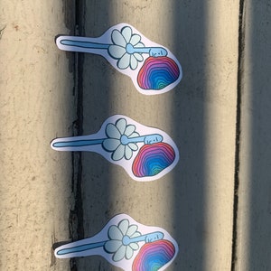 May include: Three colorful stickers with a daisy and a rainbow-colored rock with a face on it. The stickers are on a light brown wooden surface.