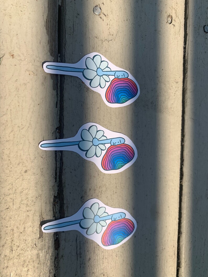 May include: Three colorful stickers with a daisy and a rainbow-colored rock with a face on it. The stickers are on a light brown wooden surface.