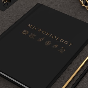 Peut inclure: Un carnet noir avec le mot "MICROBIOLOGIE" en lettres dorées sur la couverture. Le carnet est entouré de trombones et de crayons dorés sur une surface gris foncé.