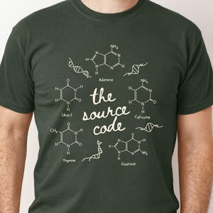 May include: Dark green t-shirt featuring a white graphic of DNA molecules and chemical structures. The text "the source code" is centered within the design. The shirt is made of a soft material.
