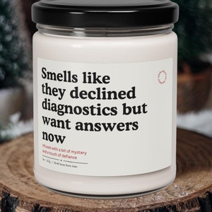 May include: A white candle with a black lid and a humorous saying: "Smells like they declined diagnostics but want answers now". The candle is on a wooden surface.